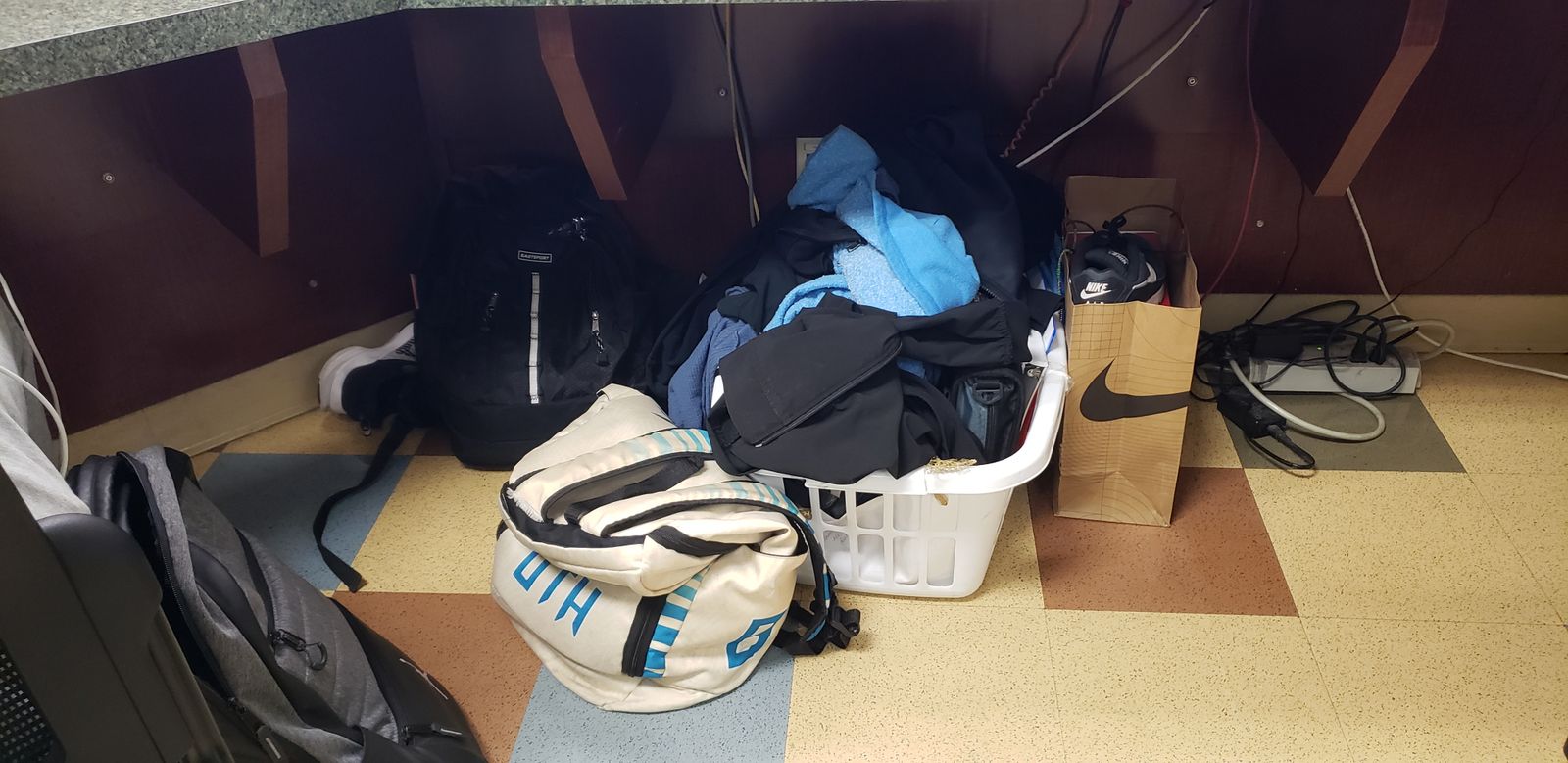 A laundry basket filled with sweatshirts sits on a floor under a desk. Next to the basket is a bag with a shoe and a backpack.