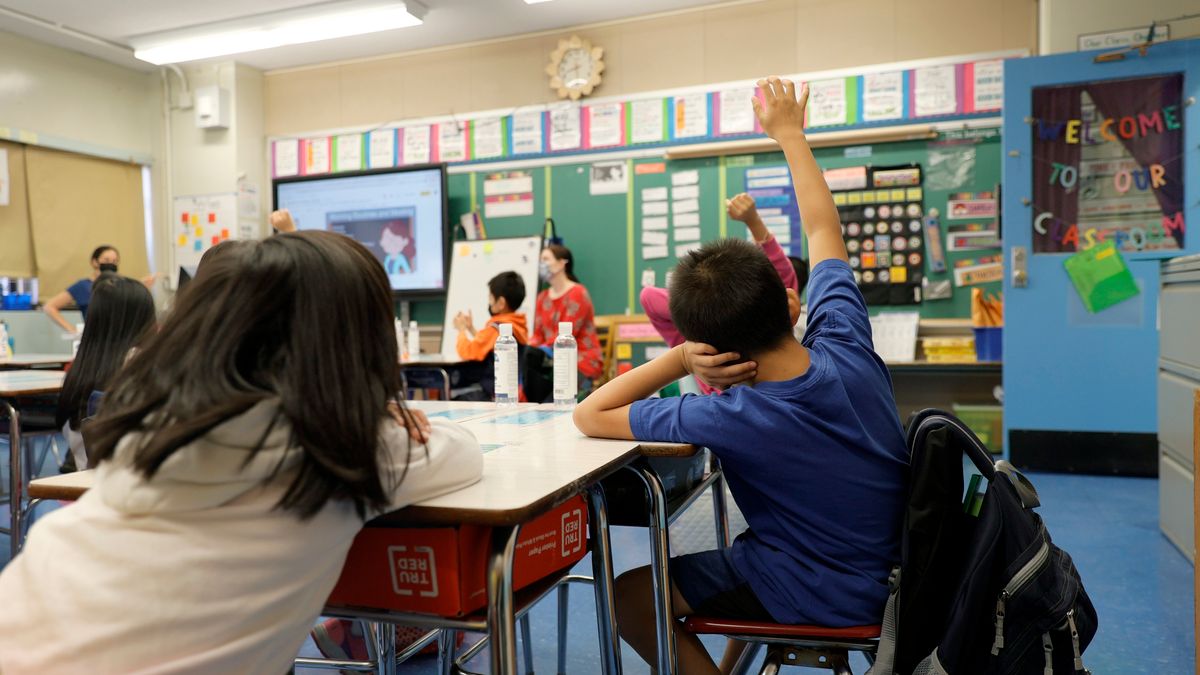 A boy and girl are sitting in the back of an elementary classroom during a lesson. One of the children is raising his hand.