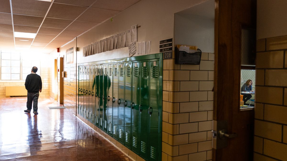A person walks down a school hallway lined with lockers as another person sitting in a classroom can be seen through a window.