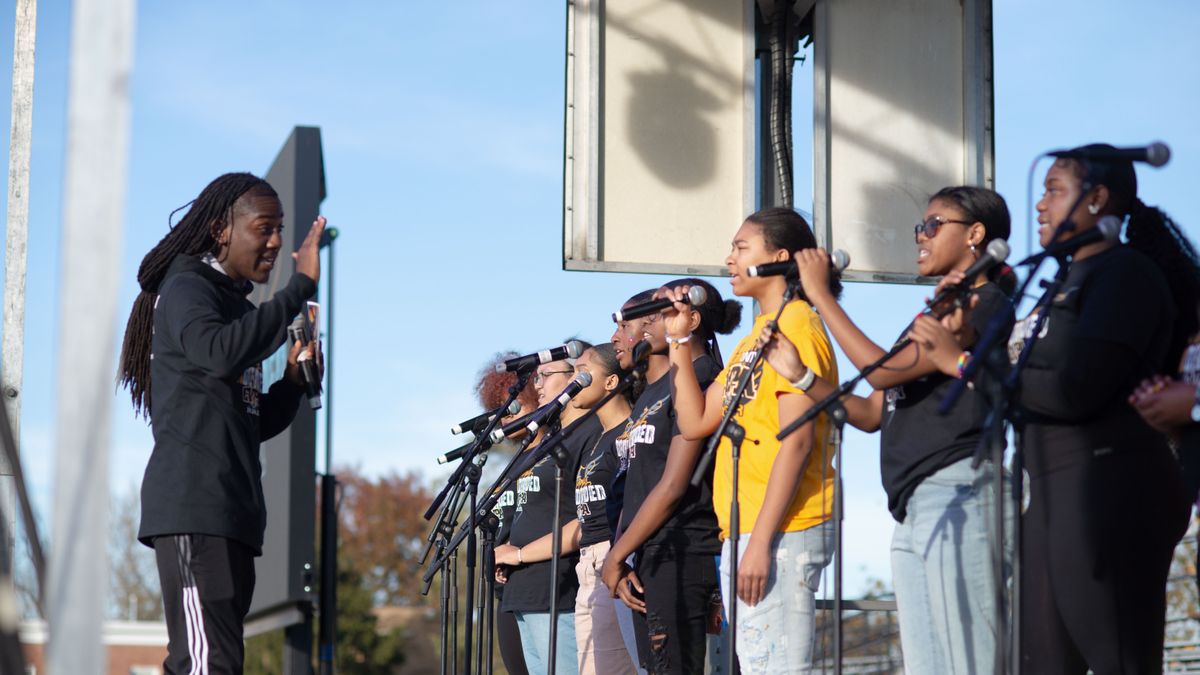 Students, standing in a row, hold microphones while singing on a stage outside.