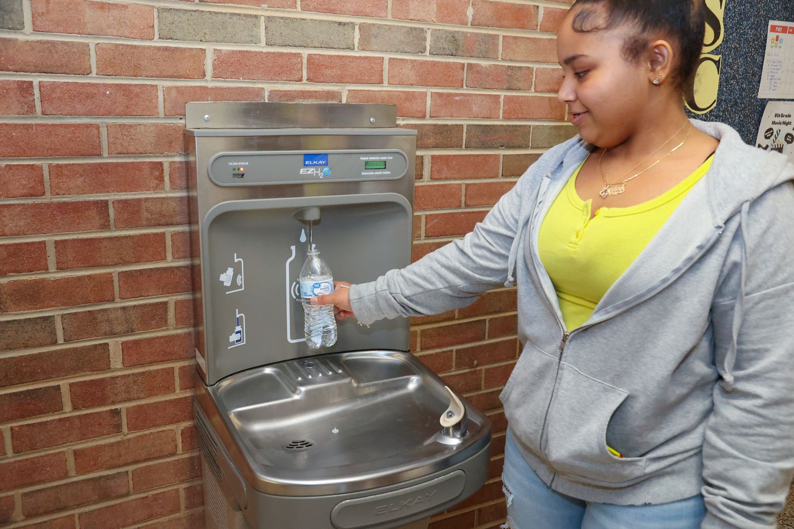 A student stands near a water fountain that has a spout for water bottles. The student is filling a bottle with water. The fountain i against a brick wall.