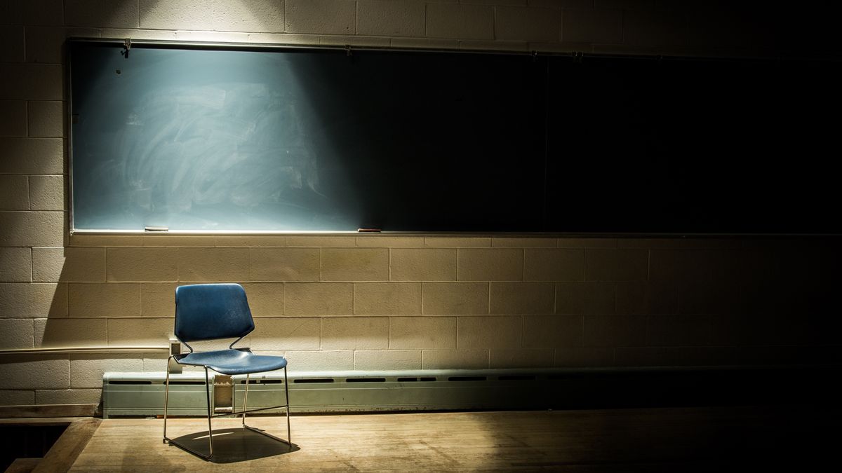 An empty chair sits in the front of a classroom. Behind the chair is a blackboard and the chair has a spotlight over it in the dark room.