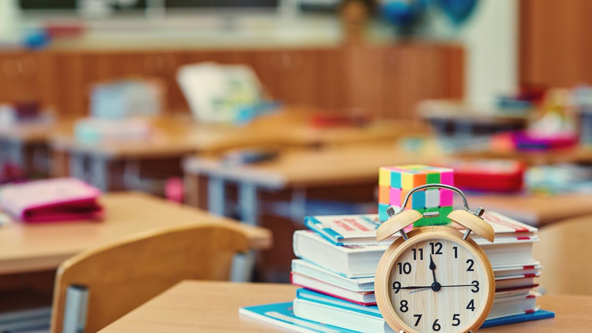 A face clock sits next to a pile of books on a stand alone desk in a classroom.