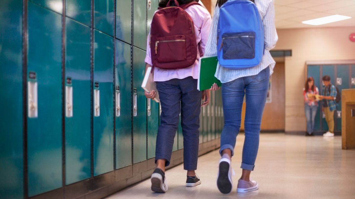 Two middle school students walk down a school hallway next to a row of lockers, backs toward the camera.