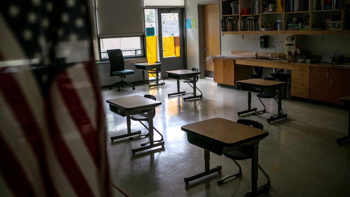 A U.S. flag hangs in the foreground of a classroom that sits empty with five visible, forlorn desks touched only by cascading rays of sunlight cutting through the dim room.