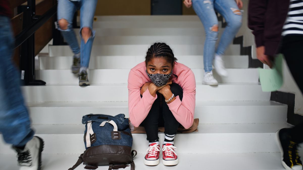 A student wearing a face mask sits on the steps in a school building staring ahead as other students walk around.