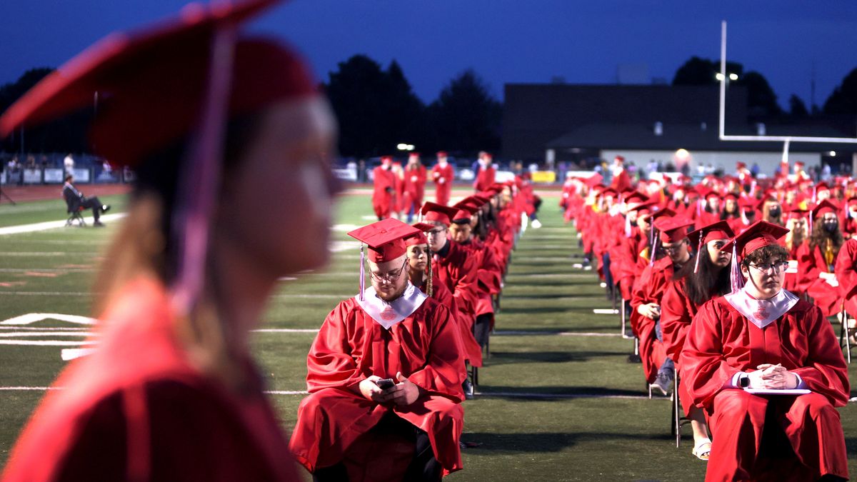 A close-up photo of a student wearing a cap and gown looking toward the side. Behind the student are rows of graduating students sitting in chairs on a football field.