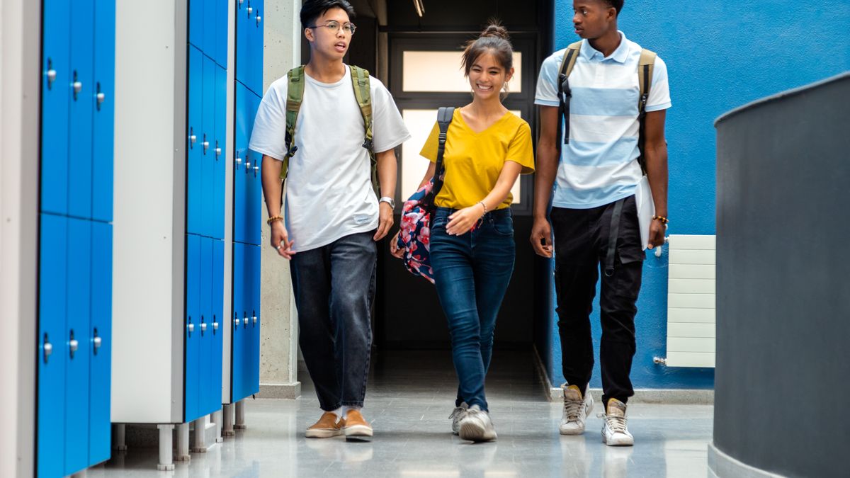 Three high school students walk down a school hallway with locker on the left side wall.