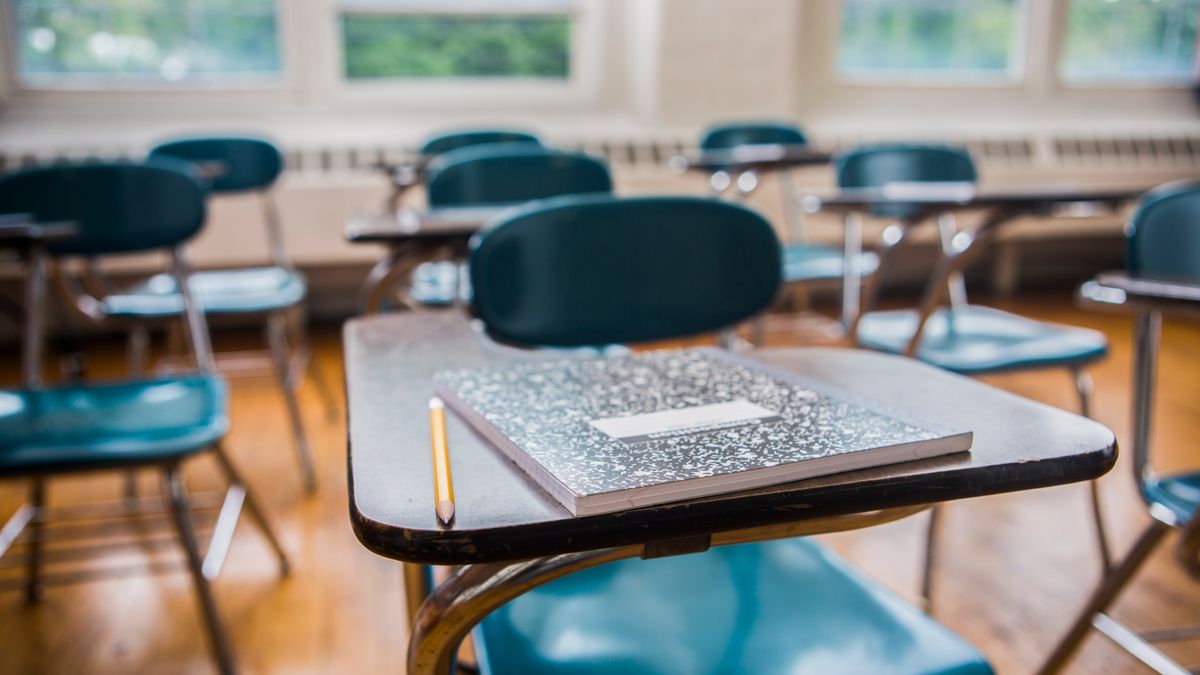 A row of desks sit empty in a classroom with the desk in the forefront having a notebook and pencil sitting on top.