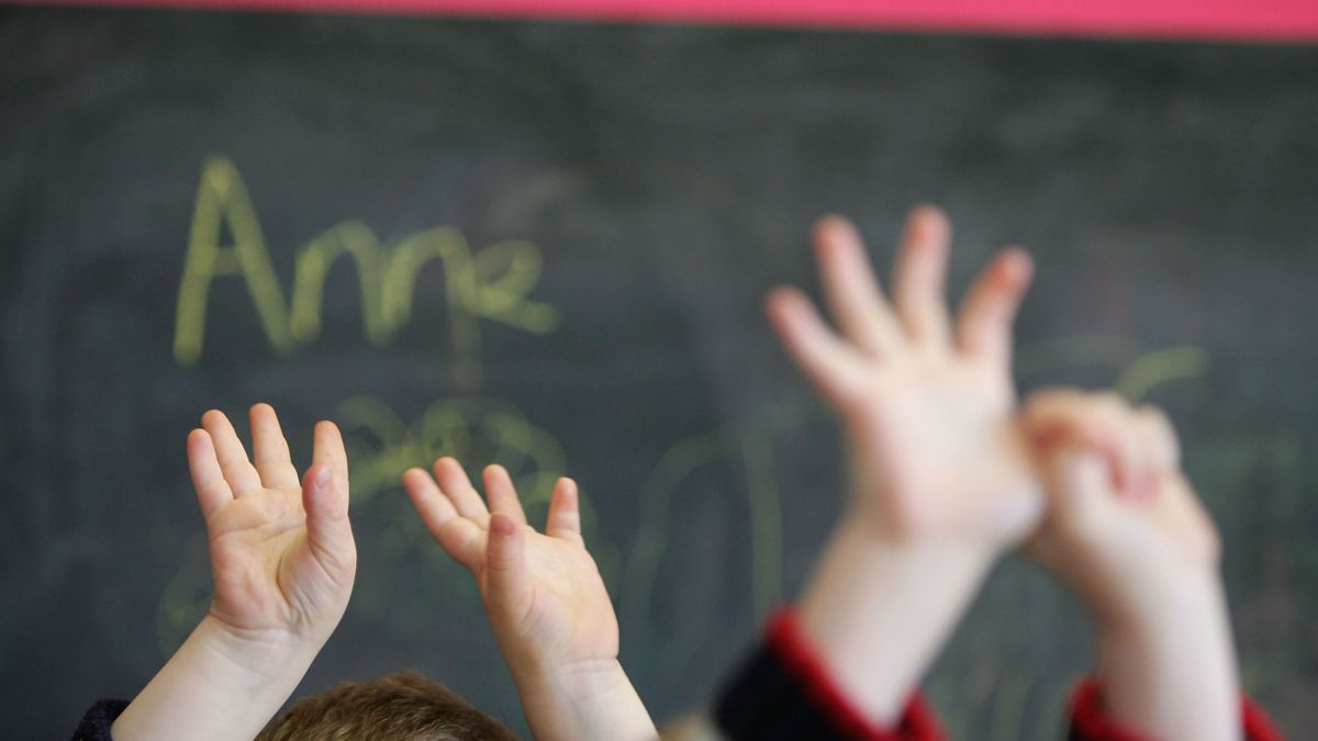 The hands of young children in a classroom are held up with a chalkboard in the background.