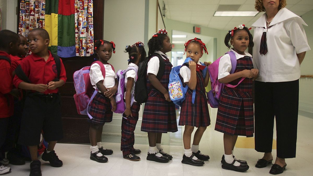 A group of Black elementary school children stand in line to the left of their teacher in a hallway.
