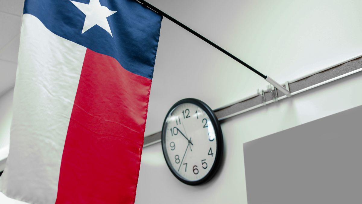 A Texas state flag hangs on a post in front of a face clock hanging in a classroom. The closeup photo shot is of the flag and clock.