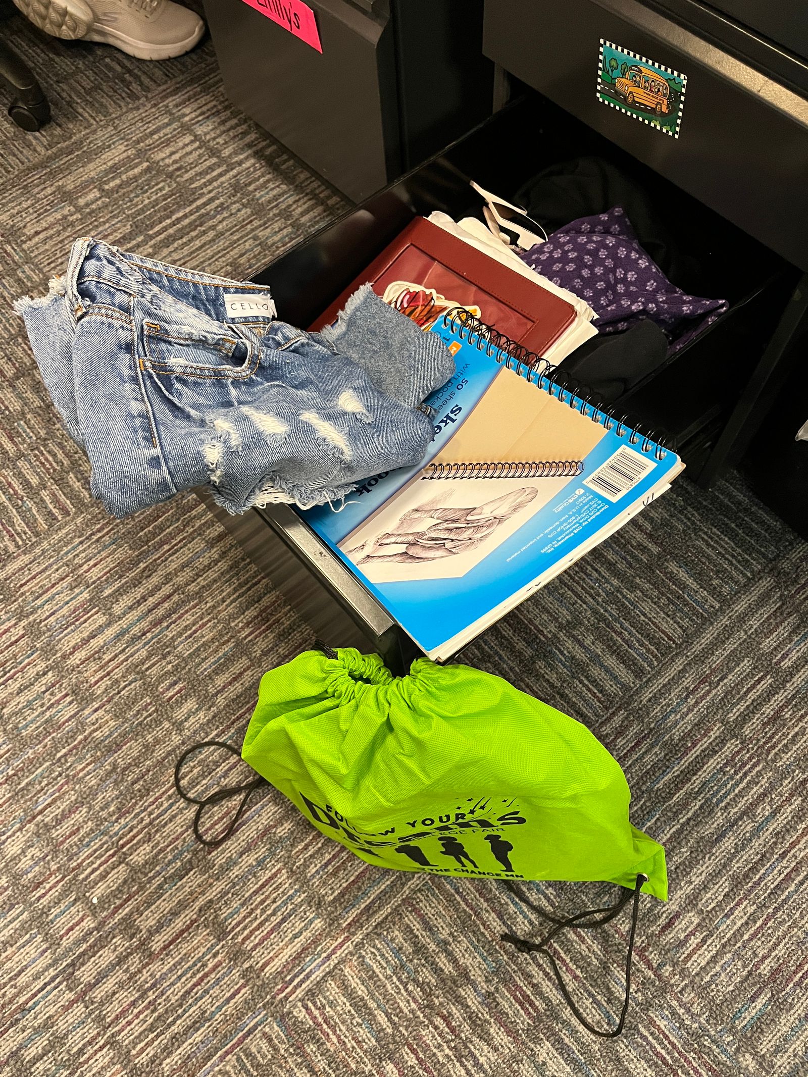 A desk drawer sits open near the floor. in the drawer are pants and notebooks. Next to the drawer on the floor is a neon green drawstring bag.