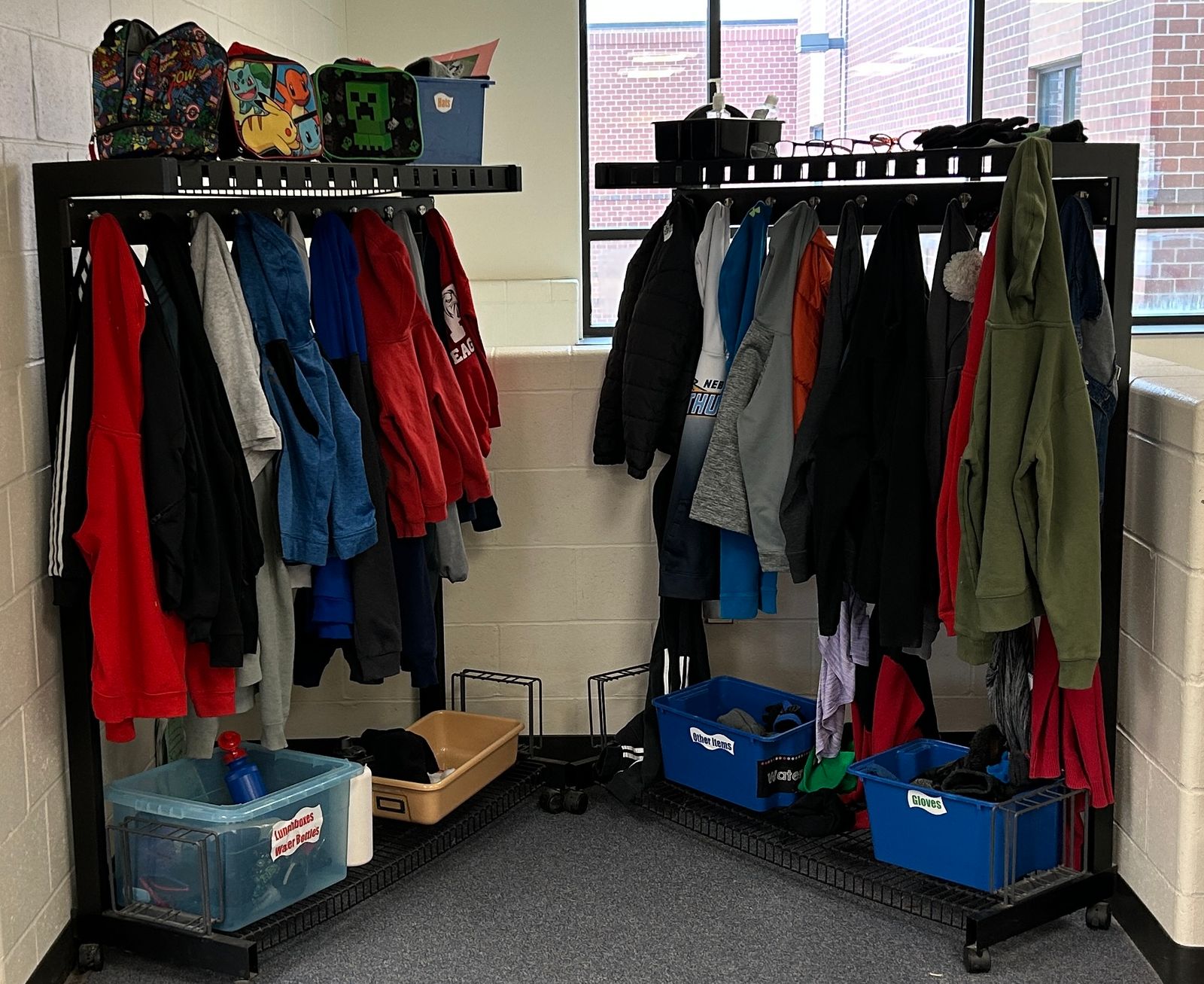 Two racks of clothes stand against walls in a school. On the floor under the racks are bins and on top of the racks are lunch boxes, glasses and other items.
