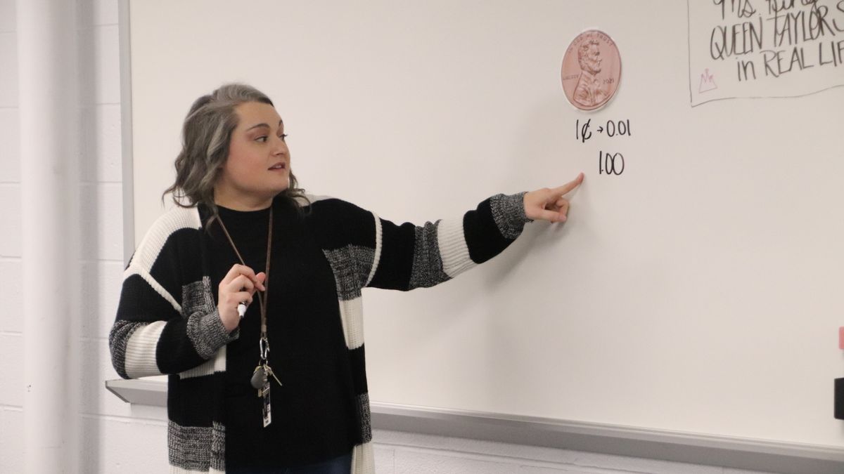 An adult stands in front of a classroom whiteboard and points to the board.