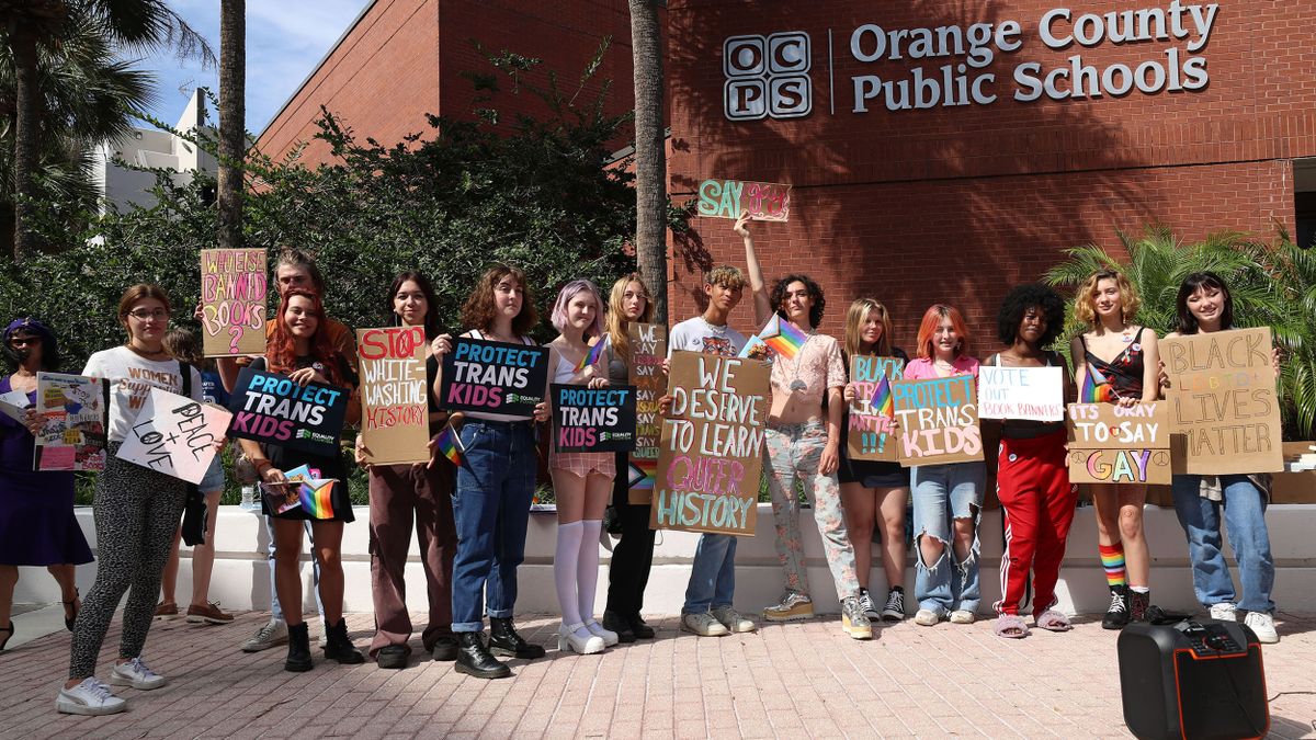 Several students hold signs that read "protect trans kids," "we deserve to learn queer history," and "vote out book banners" in front of a brick building.