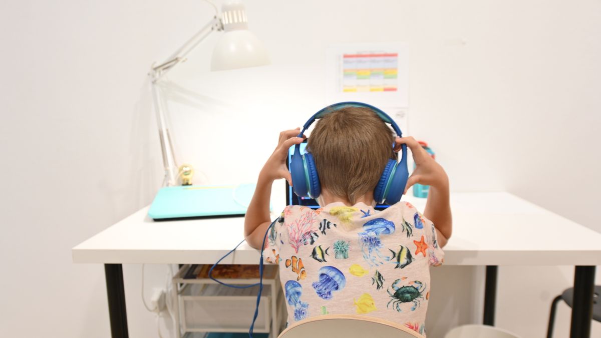 A young student has their back to the camera and is wearing headphones and sitting at a desk. A computer is on the desk.