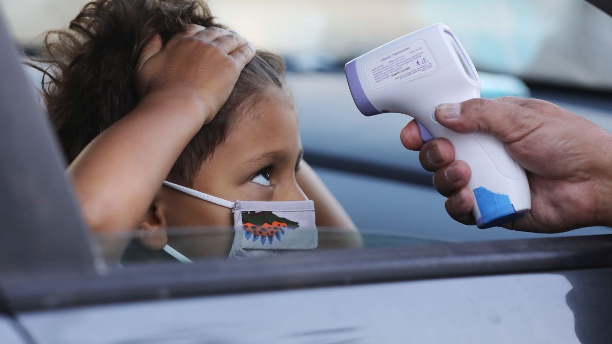 A young student sits in a car with the window down while they hold their hair back with their hands to receive a temperature check from a thermometer from someone outside of the car.