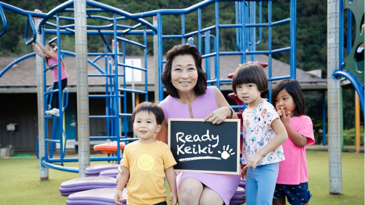 Adult is seated near an outside playground surrounded by three young children. The adult is holding a chalkboard that says "Ready Keiki"