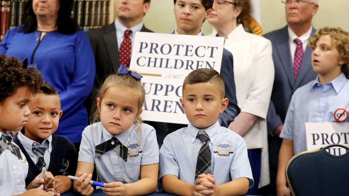 Children in uniforms congregate at a table with protest signs behind them